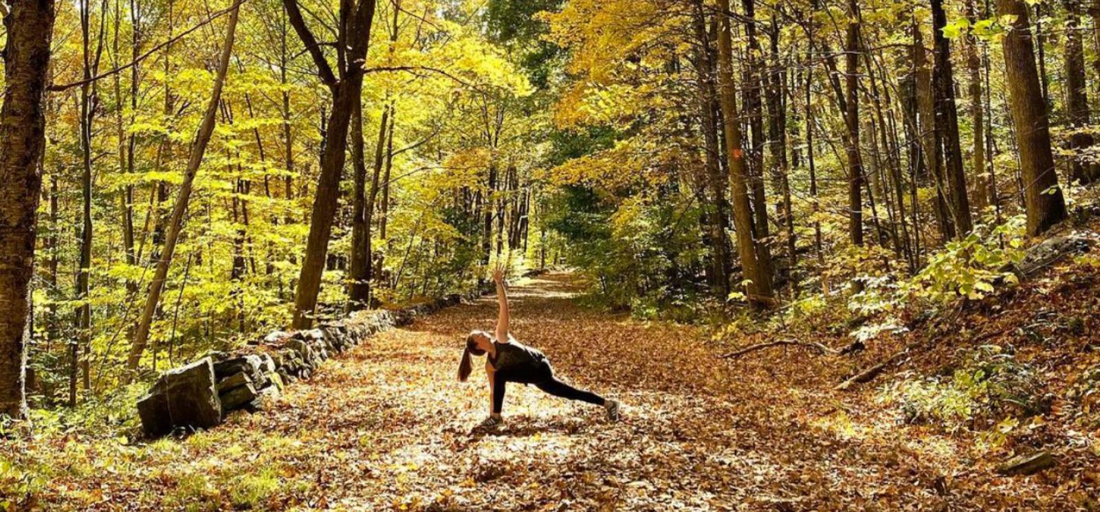 A woman doing yoga on a leaf covered path in the woods (Instagram@cjb__yogi)