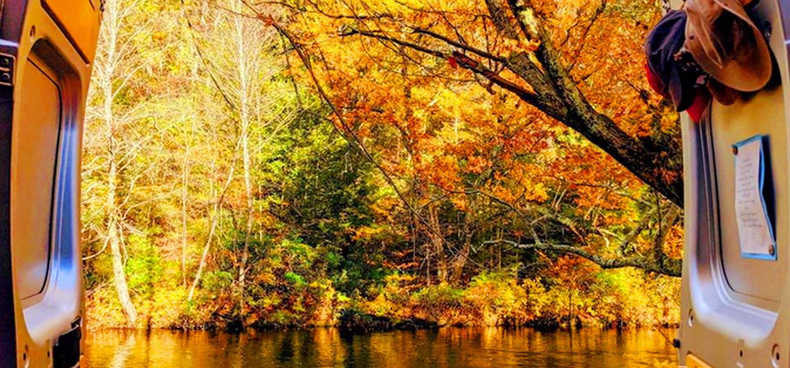 Un río que fluye entre árboles en otoño, visto a través de las puertas traseras de una camioneta.