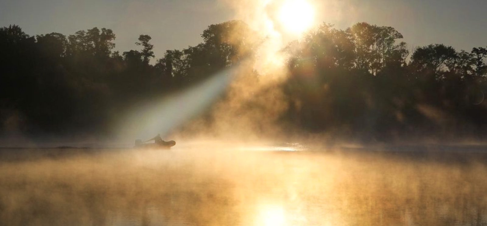 A boat glides on water as the sun pierces through the fog at River Highlands State Park.