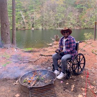 A man in a wheelchair next to a fire at a campsite in the woods (Instagram@breslifexo)