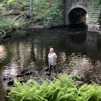 A girl stands by the water near a stone tunnel (Instagram@joeythetree)