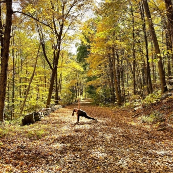 Una mujer haciendo yoga en un camino cubierto de hojas en el bosque (Instagram@cjb__yogi)