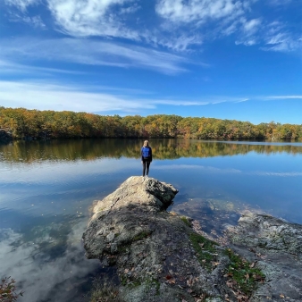 una mujer parada sobre una roca frente al agua que refleja los árboles y el cielo (Instagram@lenahrystyk)