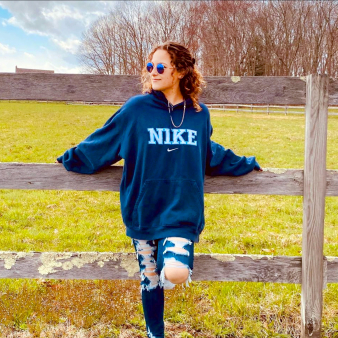 A woman in a blue sweatshirt and sunglasses leaning against a fence, enjoying the sunny day.