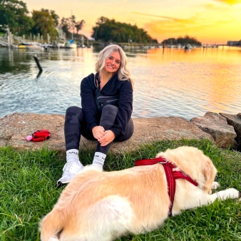 Mujer sentada en el césped junto al perro, disfrutando de una puesta de sol al aire libre.
