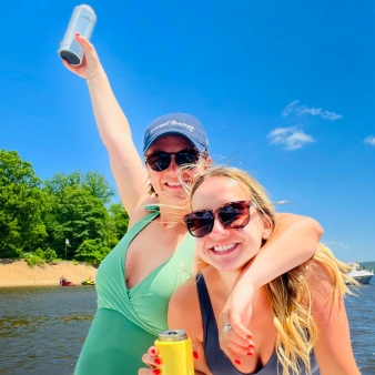 Two women in swimsuits smiling on a boat deck, enjoying a drink on a sunny day on the water.