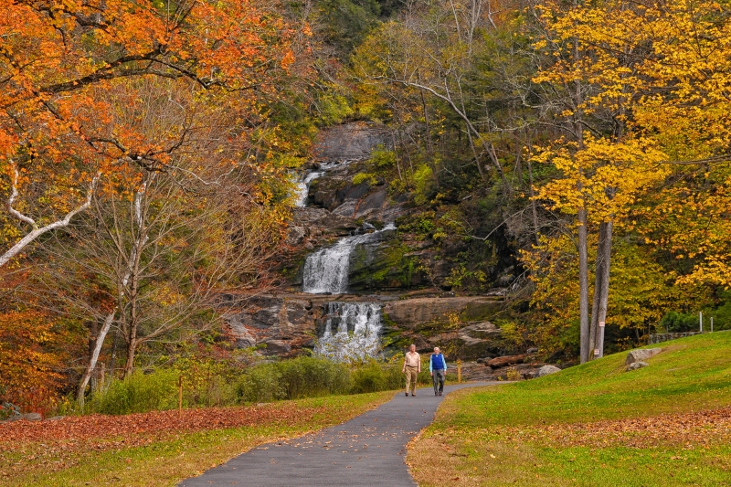 Two people walk along a trail beside a waterfall during autumn