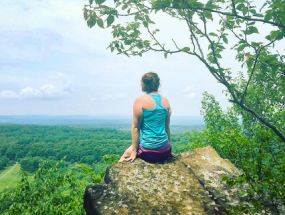 A woman peacefully sits on a rock, gazing at a breathtaking valley spread out before her.